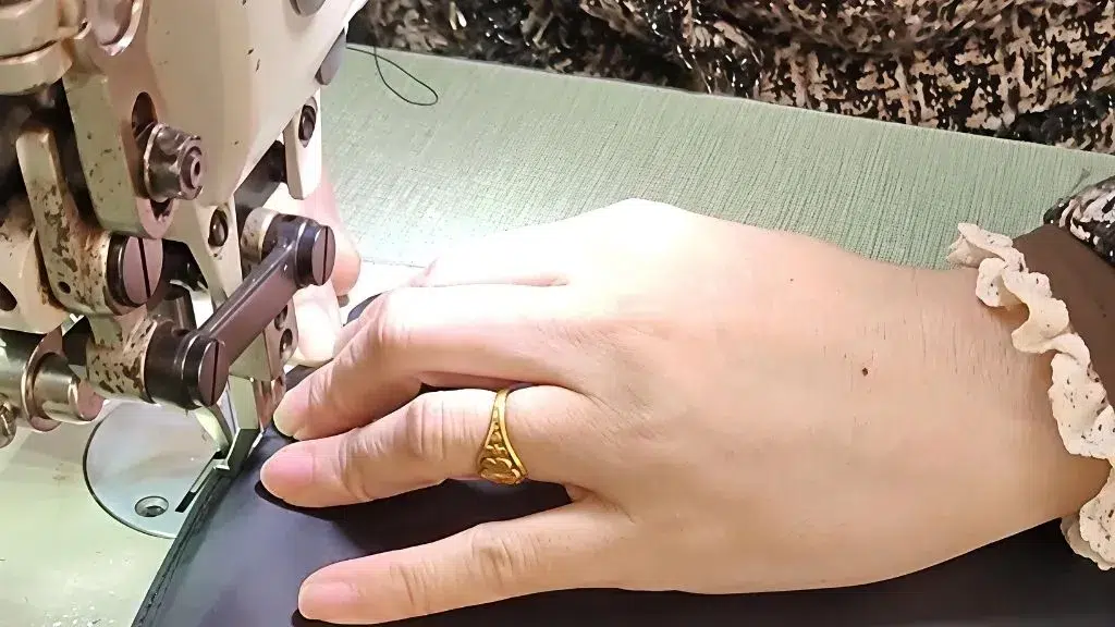 Les mains d'un artisan cousant soigneusement un sac à main en cuir marron sur une machine industrielle, montrant le processus de fabrication sur mesure.